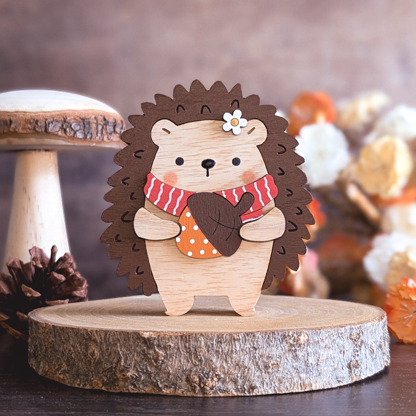 Wooden hedgehog figurine with a scarf on a wooden base, surrounded by autumn-themed decorations.
